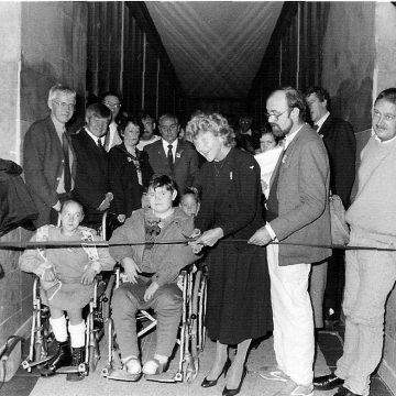 Black and white photo of a group of people on the Manchester Town Hall link bridge including two children in wheelchairs at the front. A woman cuts a ribbon across the hallway. 