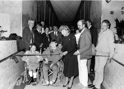 Black and white photo of a group of people on the Manchester Town Hall link bridge including two children in wheelchairs at the front. A woman cuts a ribbon across the hallway. 