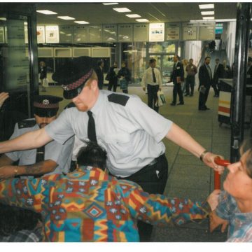 Colour photo of two police officers hold back disabled protestors at two large glass doors, with people inside the building looking on.
