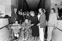 Black and white photo of a group of people on the Manchester Town Hall link bridge including two children in wheelchairs at the front. A woman cuts a ribbon across the hallway. 